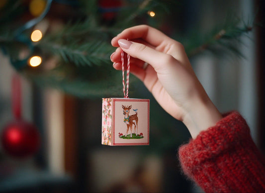 Hand holding a small Christmas ornament with a deer design in front of a decorated tree.