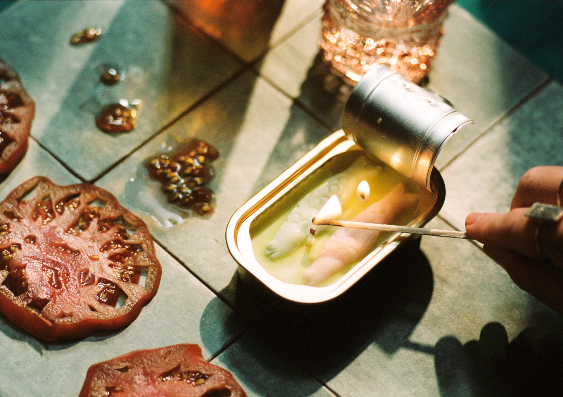 Hand lighting a candle in a metal container on a textured surface with tomatoes.